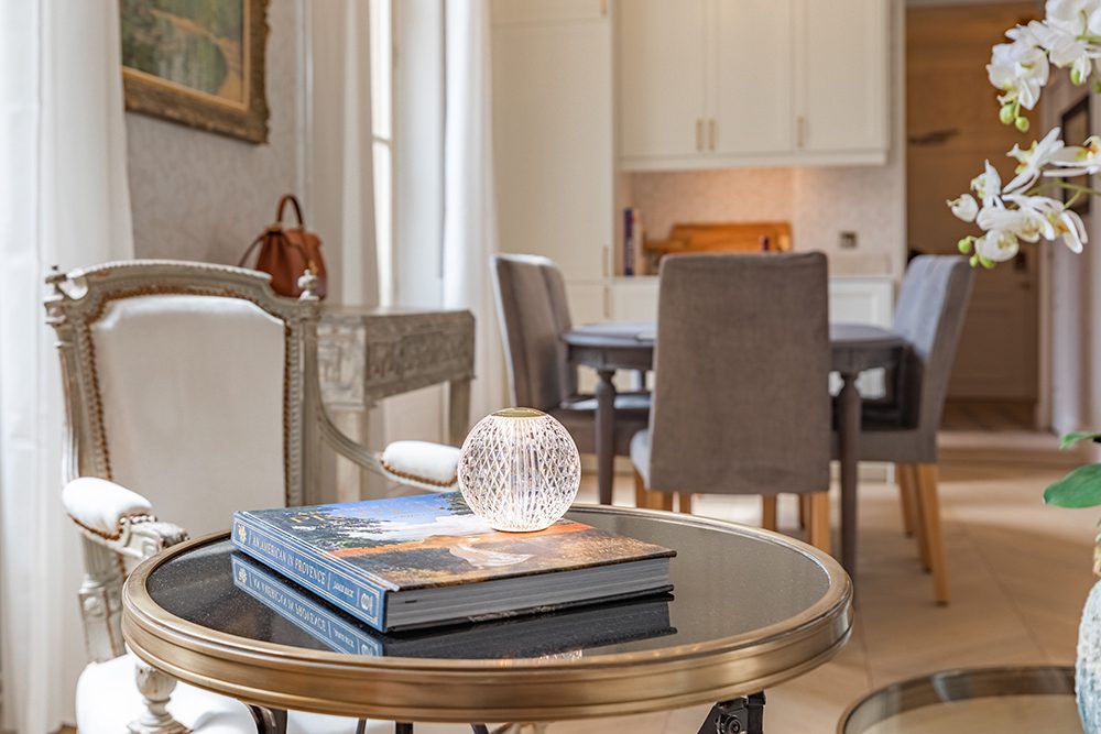 Round glass-top side table in a bright living area with a small spherical lamp and two stacked coffee-table books.